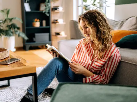 Person with curly hair sits on the floor by a couch, reading a book in a cozy living room.