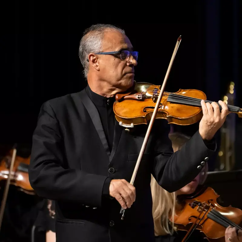 A man in a black suit plays the violin with a bow in a concert setting.