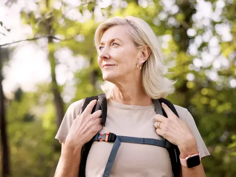 Older blonde woman wearing a backpack and smartwatch stands outdoors among sunlit green trees.
