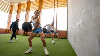 A group of young athletes in matching blue and orange uniforms performing weighted ball exercises in a modern indoor training facility with wooden architectural features and green turf flooring