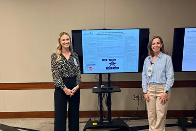 Two women stand beside a digital research poster displayed on a monitor during a conference or presentation session