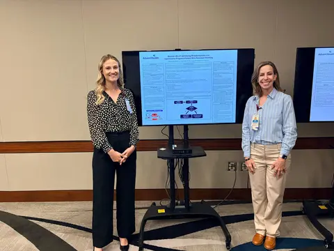 Two women stand beside a digital research poster displayed on a monitor during a conference or presentation session