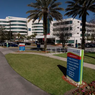 AdventHealth hospital exterior with palm trees, parking lot, and emergency sign directing to various departments.