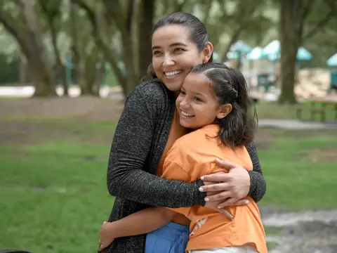 A smiling woman hugs a young girl in a park, both wearing casual clothing.