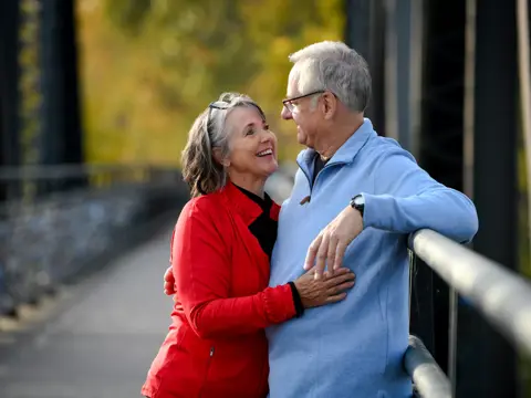Elderly couple smiling and embracing on a bridge, showcasing warmth and connection.