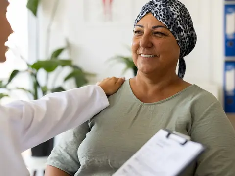 Doctor in white coat places a reassuring hand on the shoulder of a smiling middle-aged female patient wearing a patterned headscarf in a bright medical office