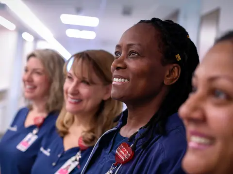 Four smiling nurses in blue uniforms stand together in a hospital corridor, embodying warmth and care.