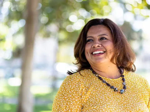 Person outdoors smiles while looking to the side, wearing a yellow patterned top and necklace.