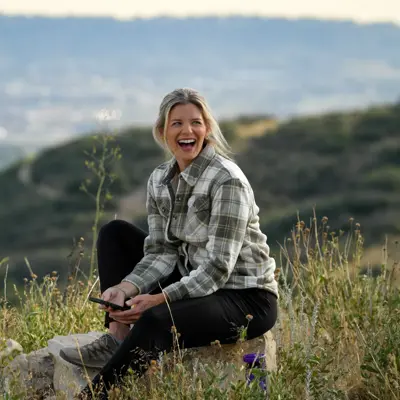 A woman sits on a rock, smiling and holding a smartphone, with a scenic view in the background.