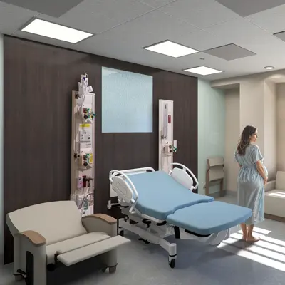 Pregnant woman in a hospital gown standing by a large window overlooking trees in a modern maternity room with a hospital bed and seating area