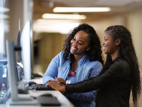 Two smiling nurses in blue scrubs working together at a computer workstation in a hospital setting