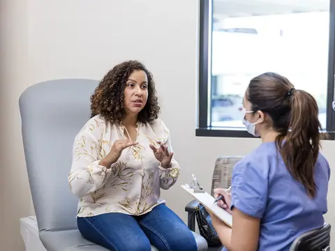 Seated woman gestures while speaking with a masked person in scrubs taking notes on a clipboard.