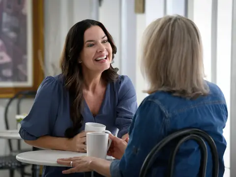 Two women sitting at a table having a warm conversation over coffee in a bright, naturally lit room.