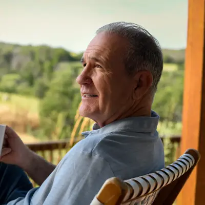 An older man sitting on a porch, holding a coffee mug, and looking out at a scenic view.