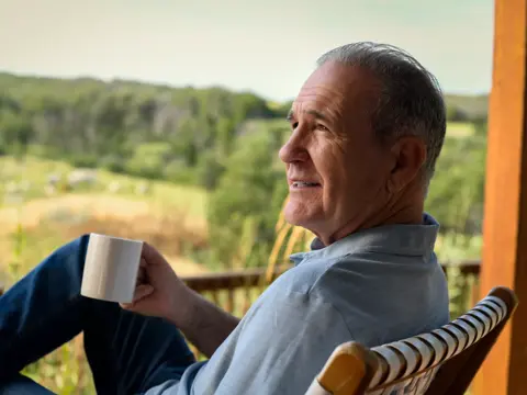 An older man sitting on a porch, holding a coffee mug, and looking out at a scenic view.