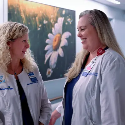 Two smiling nurses in white coats stand in a hospital corridor, one wearing a blue shirt.
