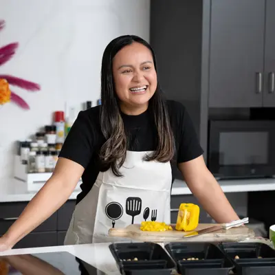 A smiling woman in a kitchen, wearing an apron with cooking tools, preparing food.