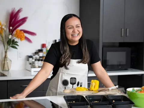 A smiling woman in a kitchen, wearing an apron with cooking tools, preparing food.