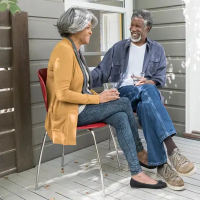Two people sit on a porch, smiling at each other and holding glasses, with sunlight and shadows across the deck.
