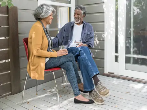 Two people sit on a porch, smiling at each other and holding glasses, with sunlight and shadows across the deck.