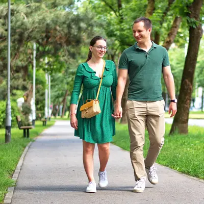 Two people hold hands and smile while walking on a tree-lined park path, with benches and lampposts in the background.