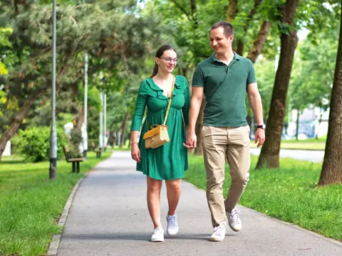 Two people hold hands and smile while walking on a tree-lined park path, with benches and lampposts in the background.