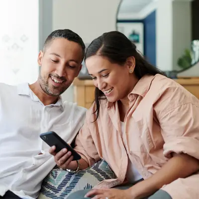 Two people sit together on a couch, smiling as they look at a smartphone in a bright living room.
