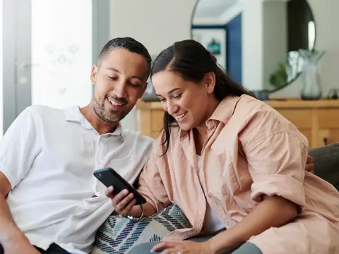 Two people sit together on a couch, smiling as they look at a smartphone in a bright living room.