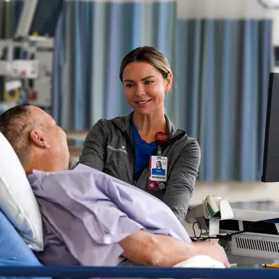 AdventHealth team member in gray jacket smiling while speaking with a male patient reclining in a hospital bed in a surgical services room with medical equipment and blue privacy curtains visible in the background