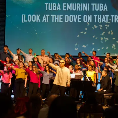 Colorfully dressed choir points outward while singing onstage beneath projected lyrics reading 'TUBA EMURINI TUBA [LOOK AT THE DOVE ON THAT TREE!]'
