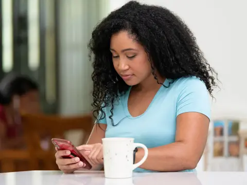 Woman in light blue shirt looking down at her smartphone while sitting at a table with a white coffee mug in a home setting