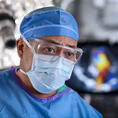 Surgeon in blue scrubs, cap, and mask, with protective eyewear, in an operating room setting.