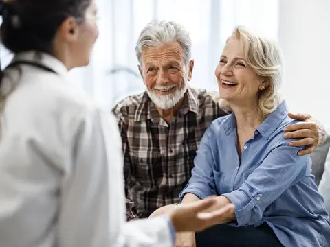 Older couple smiling while talking with a person wearing a white coat in a bright room.