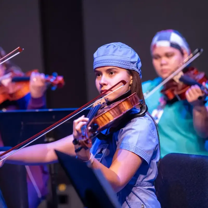 Ivanna Mirabal Molina plays viola in blue scrubs and a surgical cap alongside other string musicians.