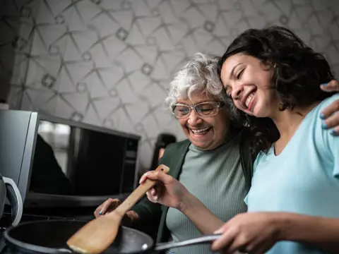 Two women cooking together in a kitchen, sharing a laugh and a hug.