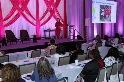 Speaker in a pink dress presenting to an audience at a women's health event, with a "Lessons I learned along the way" slide displayed on screen.