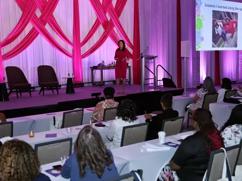 Speaker in a pink dress presenting to an audience at a women's health event, with a "Lessons I learned along the way" slide displayed on screen.
