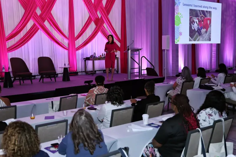 Speaker in a pink dress presenting to an audience at a women's health event, with a "Lessons I learned along the way" slide displayed on screen.