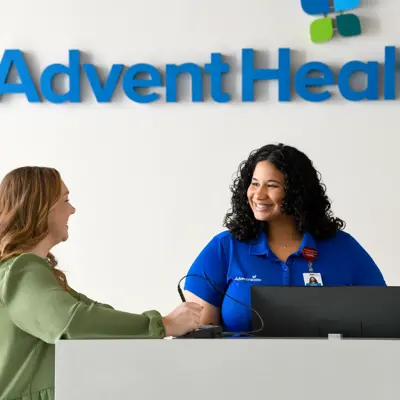 A smiling woman in a blue shirt assists a customer at the AdventHealth reception desk.