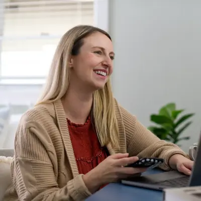A smiling woman sits at a desk with a laptop and candle, holding a remote.