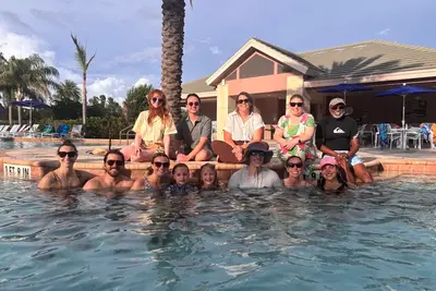 Group of adults and children gather in and around a swimming pool, smiling at the camera in front of a clubhouse and palm trees.