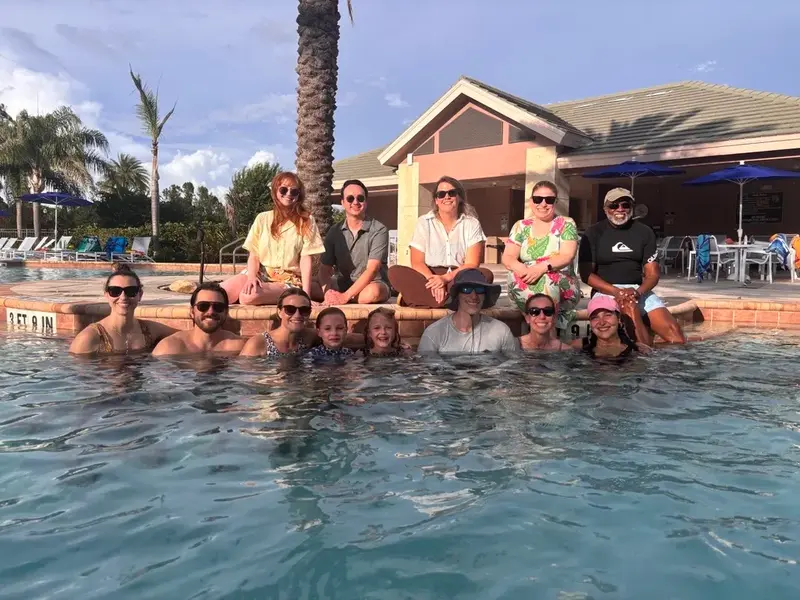 Group of adults and children gather in and around a swimming pool, smiling at the camera in front of a clubhouse and palm trees.