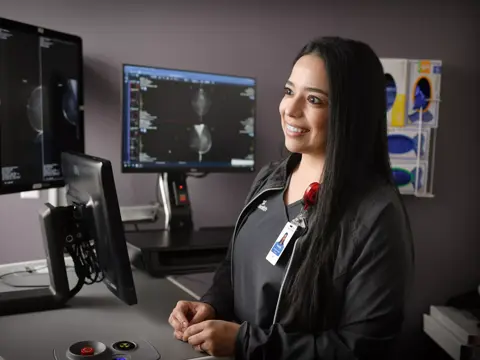 A smiling woman in a black jacket stands in front of medical monitors in a clinic.