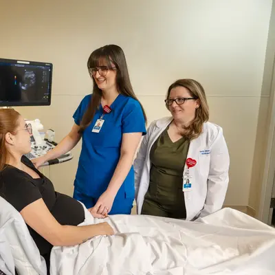 Three medical professionals in white coats and blue scrubs standing around a pregnant patient seated in an exam chair during an ultrasound consultation in a medical training room