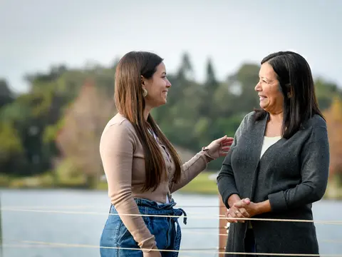 Two Hispanic women smiling and talking together outdoors near a lake with trees in the background