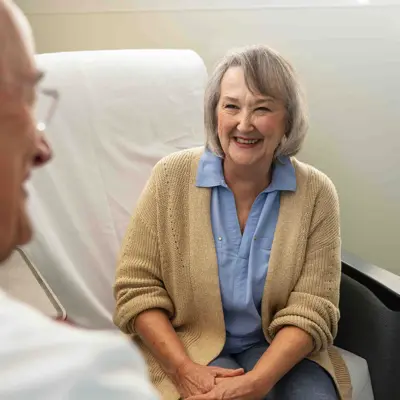 Smiling older woman patient in beige cardigan sitting across from physician in white coat during consultation in medical office