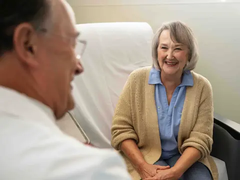 Smiling older woman patient in beige cardigan sitting across from physician in white coat during consultation in medical office