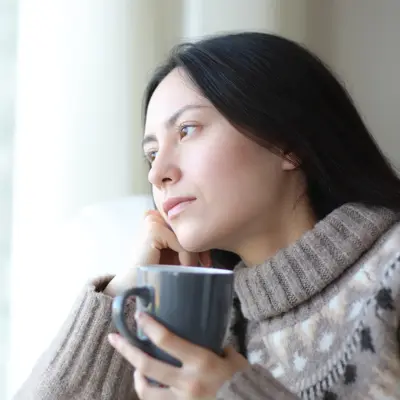 Person in a patterned turtleneck sweater holds a dark mug and gazes out a sunlit window with a thoughtful expression.
