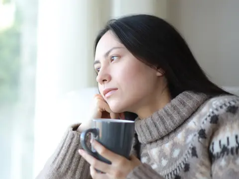 Person in a patterned turtleneck sweater holds a dark mug and gazes out a sunlit window with a thoughtful expression.