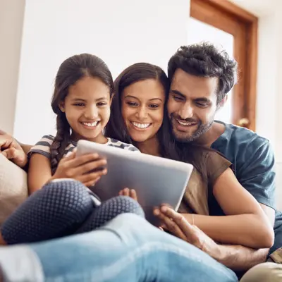 A family of three smiling and looking at a tablet together on a couch.